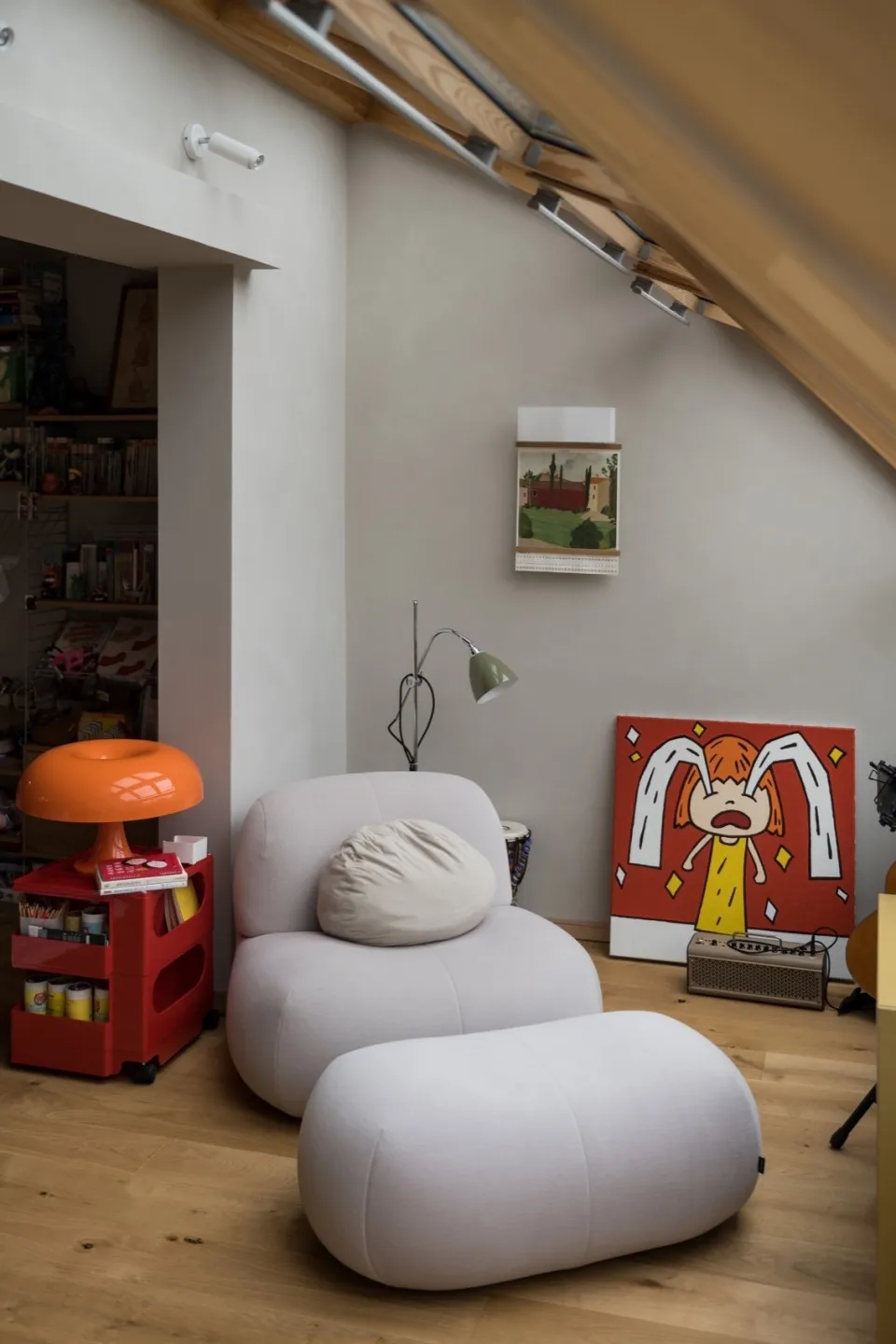 A minimalist living space featuring a white upholstered chair and ottoman, a red and orange lamp, and a red painting of a cha