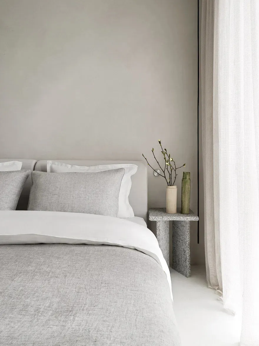 A minimalist bedroom featuring a bed with gray and white textiles, a side table with plants and vases, and white curtains.