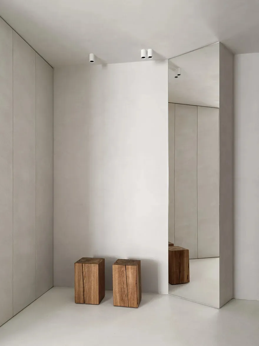 Minimalist room with large white walls, wooden stools, and a tall mirror.
