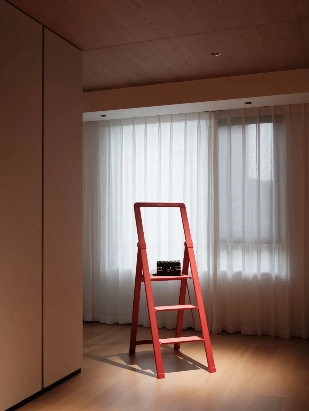 Red metal step ladder with a black purse on it positioned against a white curtain in a modern home interior.