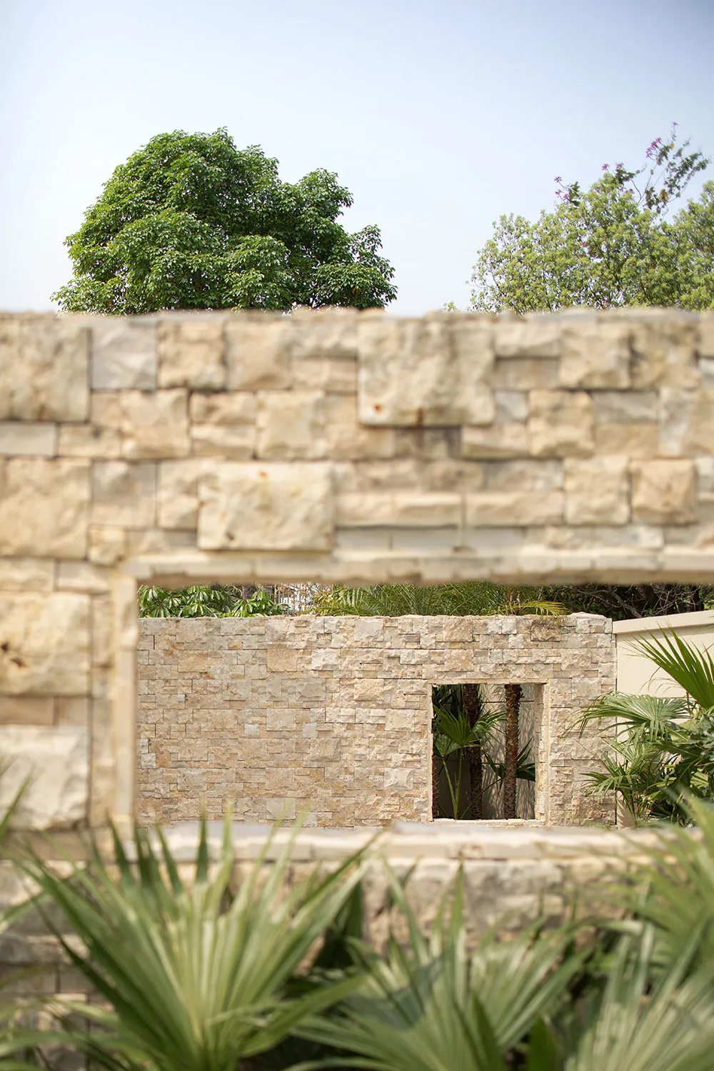 A view of a stone wall in a garden setting with green foliage and trees in the background.