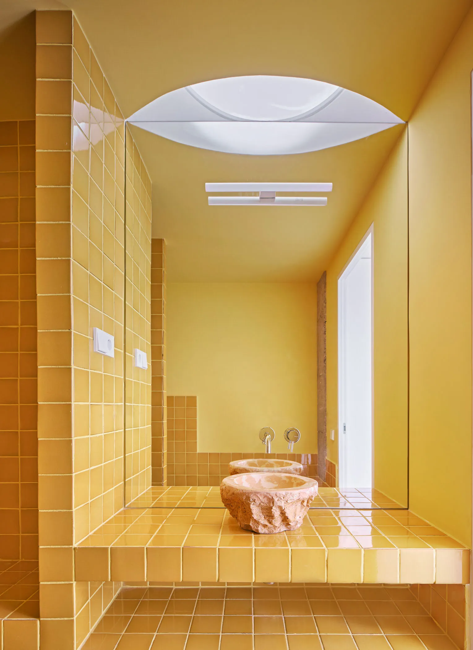 A minimalist bathroom with yellow ceramic tiles, a stone sink, and a large mirror.