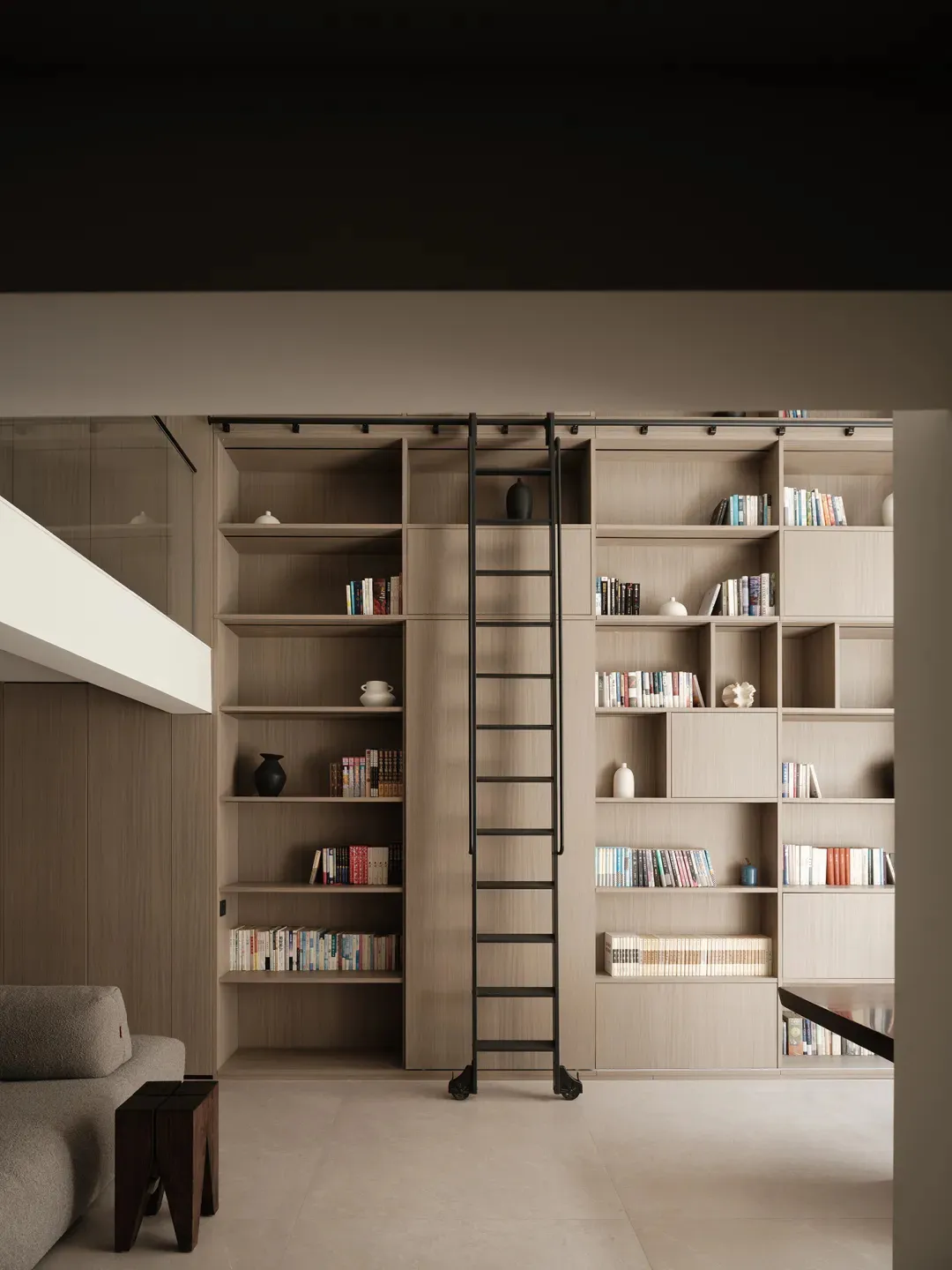 Minimalist living room with wooden bookshelves and ladder, featuring a grey couch and dark wooden stools.