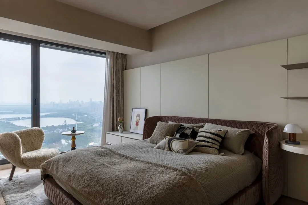 A modern bedroom with a brown velvet bed, a sleek desk, and a large window overlooking a cityscape.