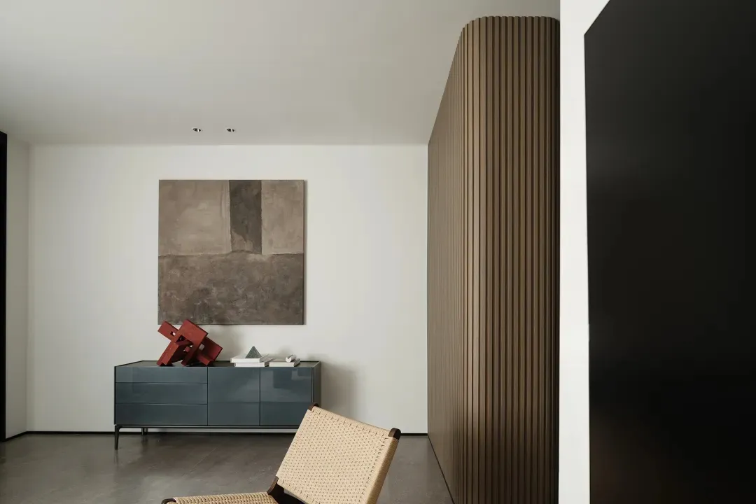 A minimalist bedroom with a modern design, featuring a wooden chair, a blue dresser, and a painting on the wall.