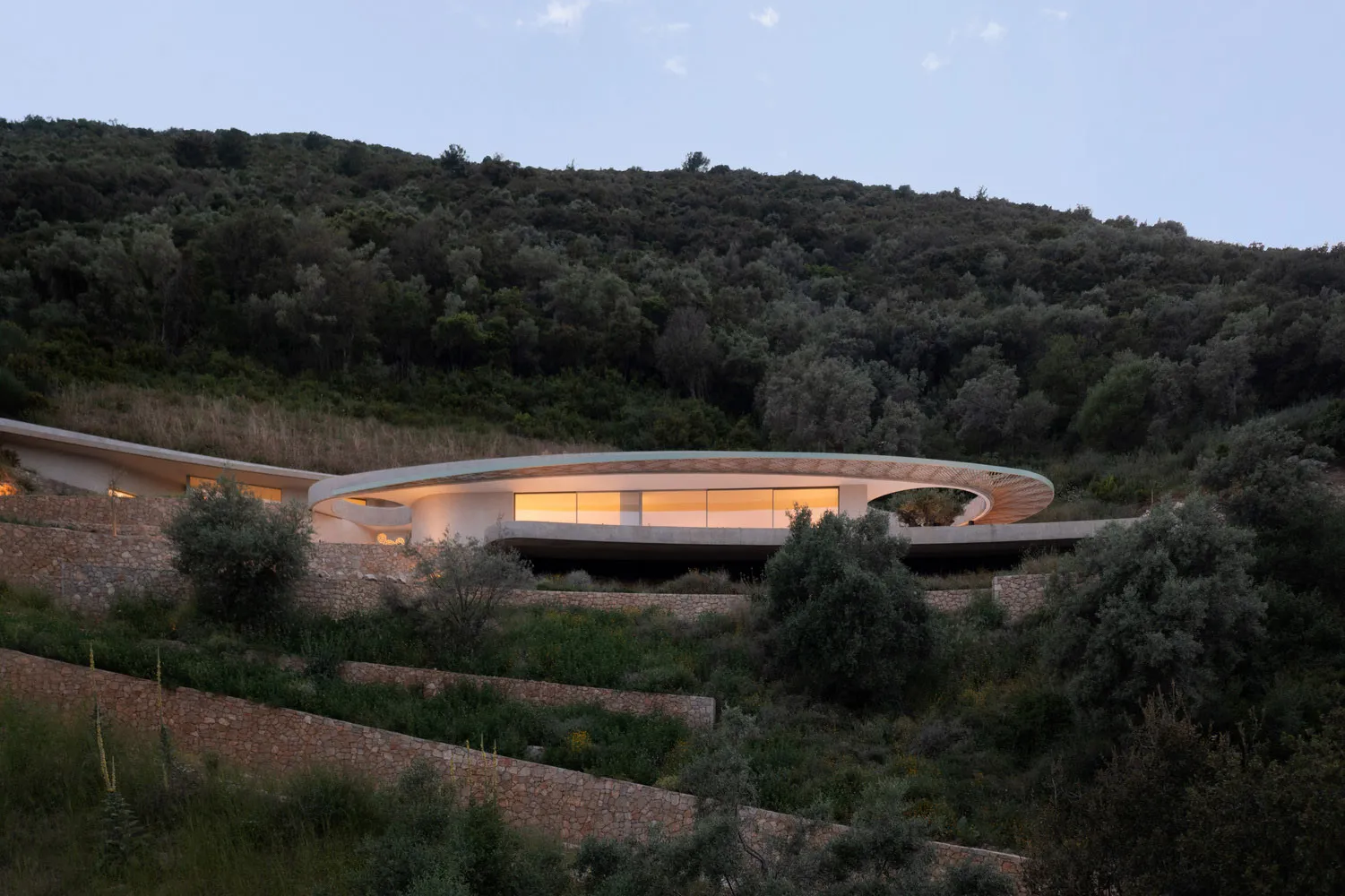 The image depicts a modern residential house with a circular design and concrete material, surrounded by a natural landscape