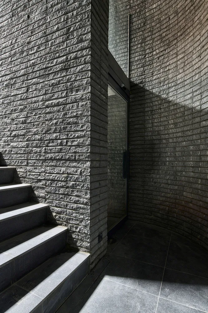 Modern entryway featuring dark gray stairs adjacent to a curved brick wall with a glass door.