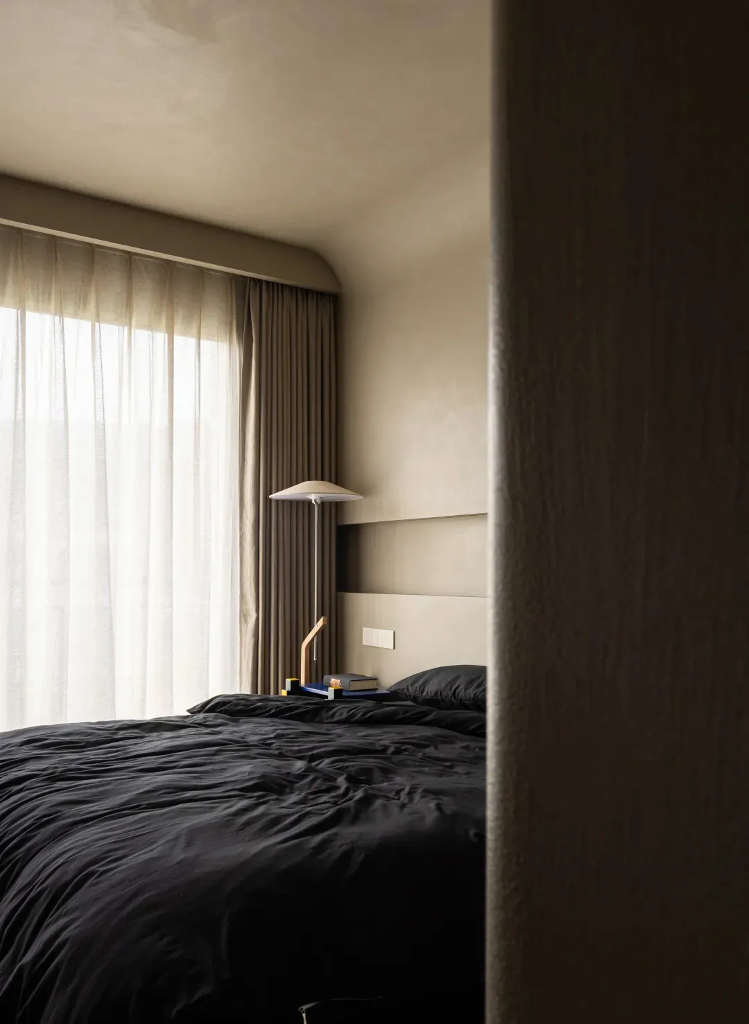 A minimalist bedroom with a textured wall, a bed with black bedding, and a lamp on a headboard.