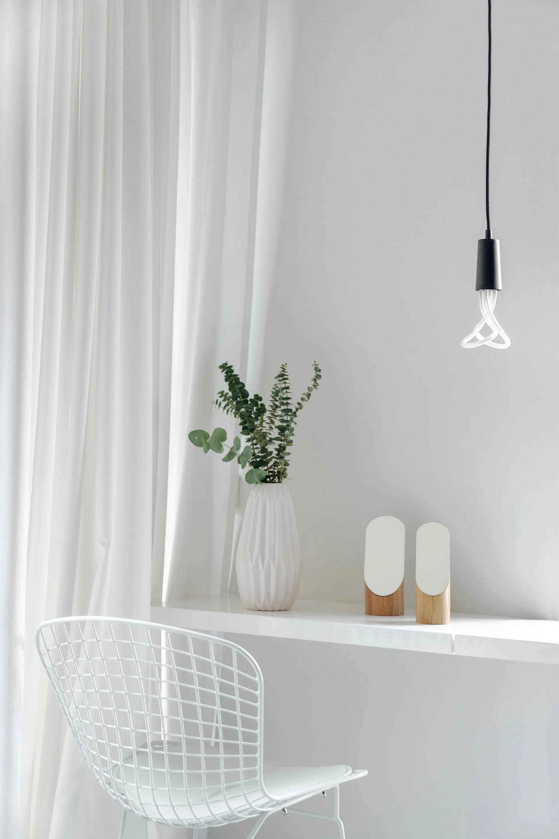 A minimalist home office with a white desk, white wire chair, green plant, and white hanging light.