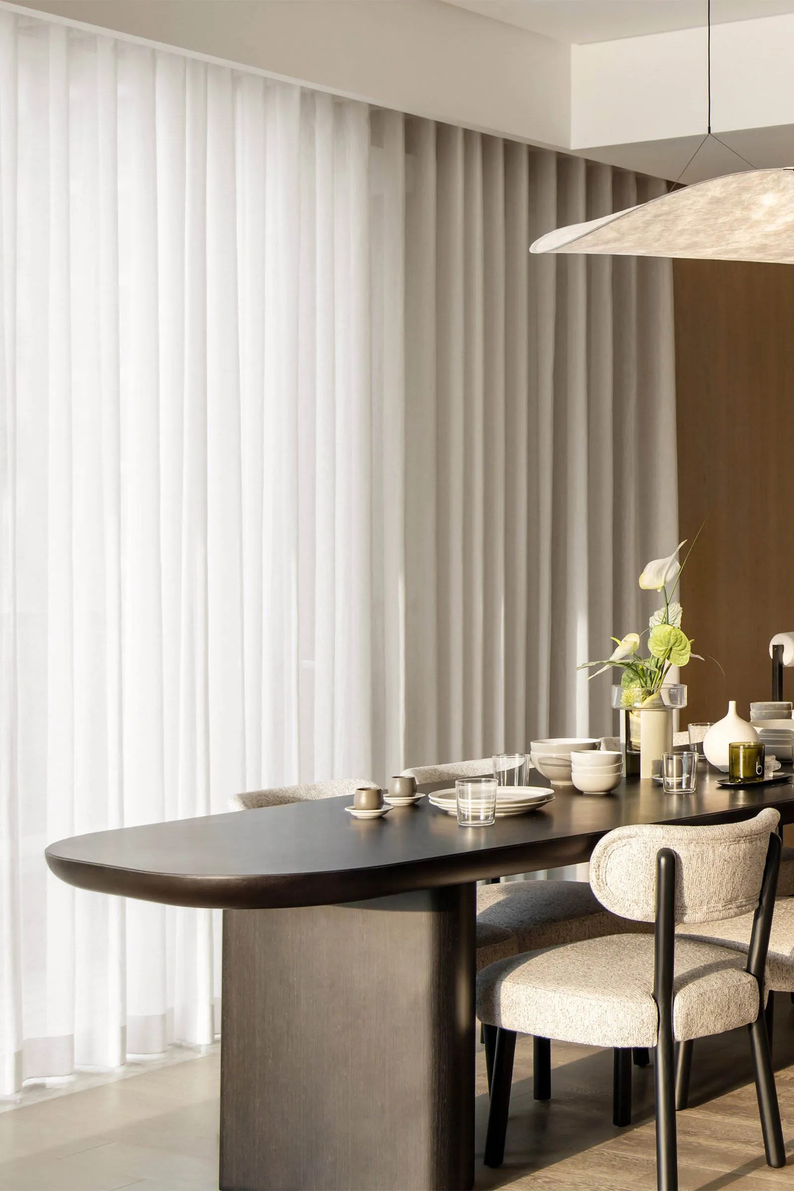 A modern minimalist dining room featuring a sleek, dark wooden table and beige upholstered chairs, surrounded by sheer curtai