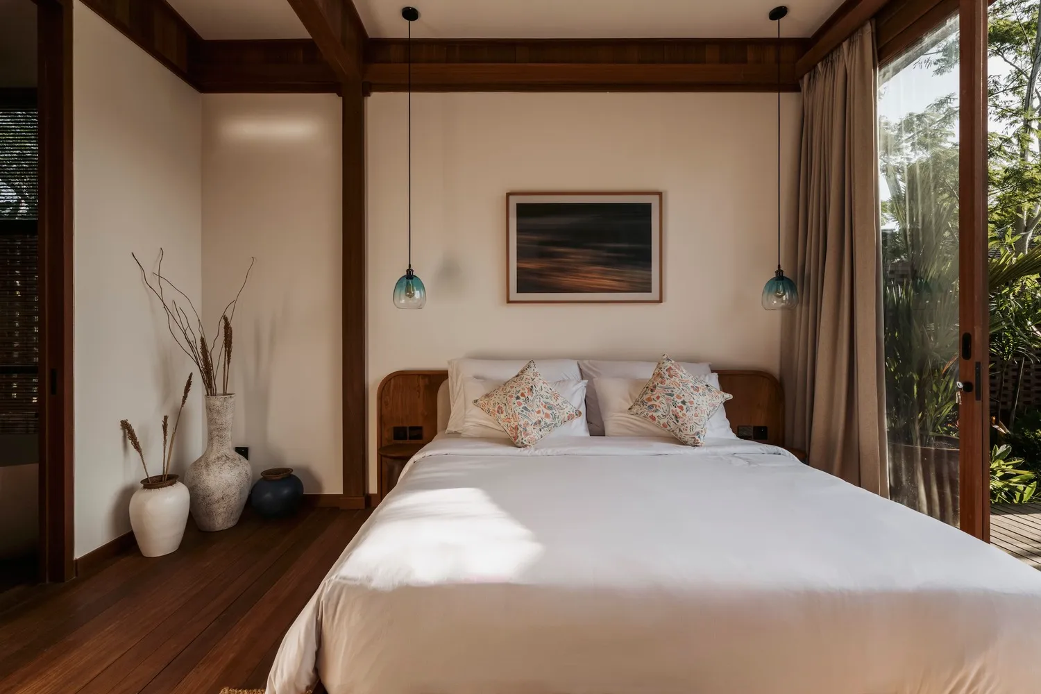 A minimalist bedroom with a wooden bed and white bedding, featuring a painting and blue pendant lights.