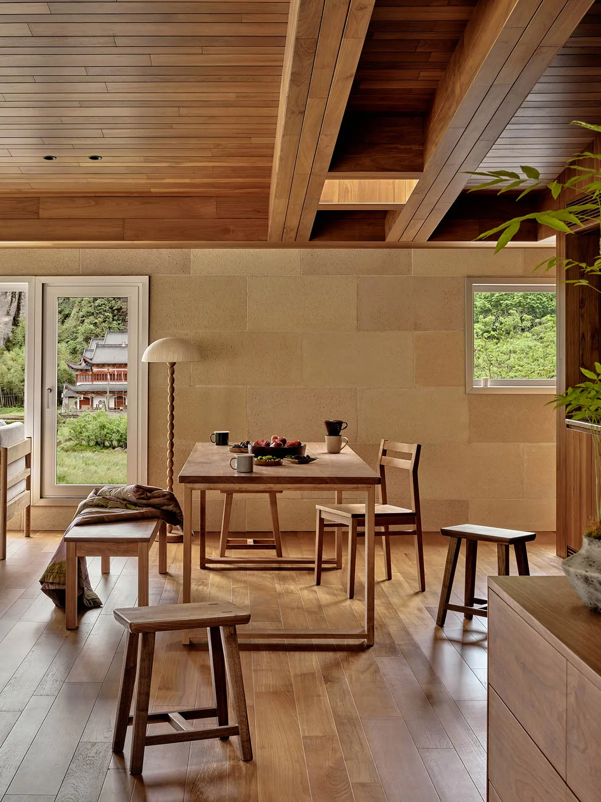 A minimalist dining room with wooden furniture and a wooden floor, featuring natural light and a stone wall.