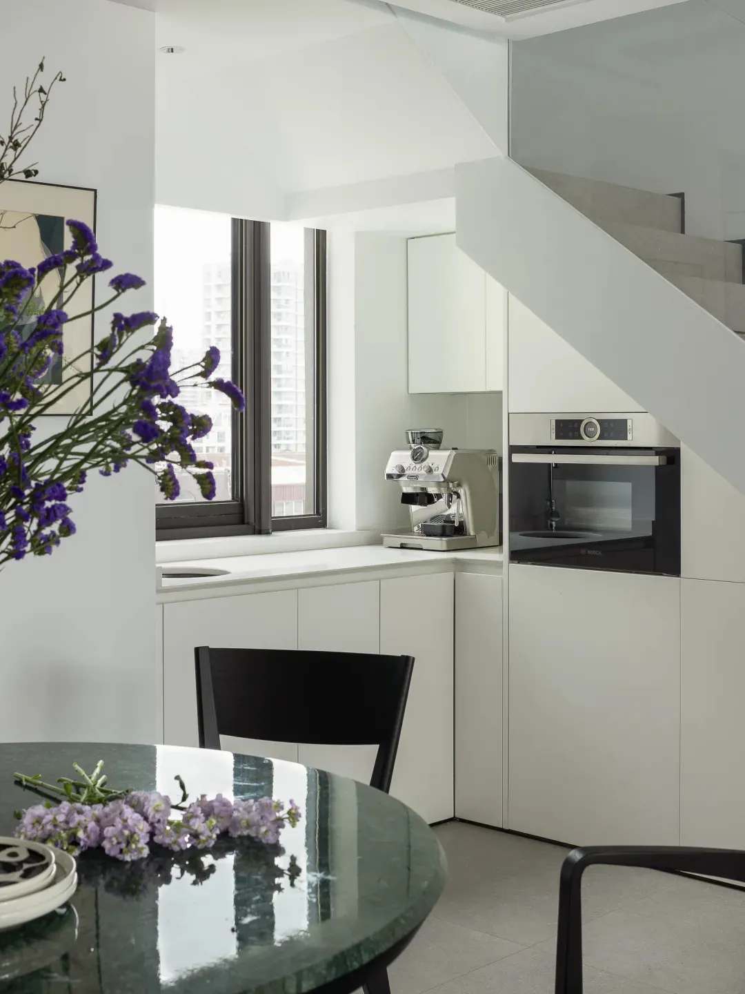 A modern kitchen featuring white cabinets, a black marble table with purple flowers, and a window with a cityscape view.