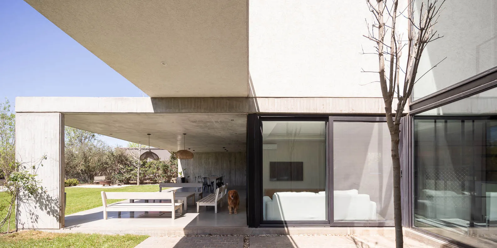 A modern residential exterior featuring a concrete structure with large glass windows and a dog standing on the porch.