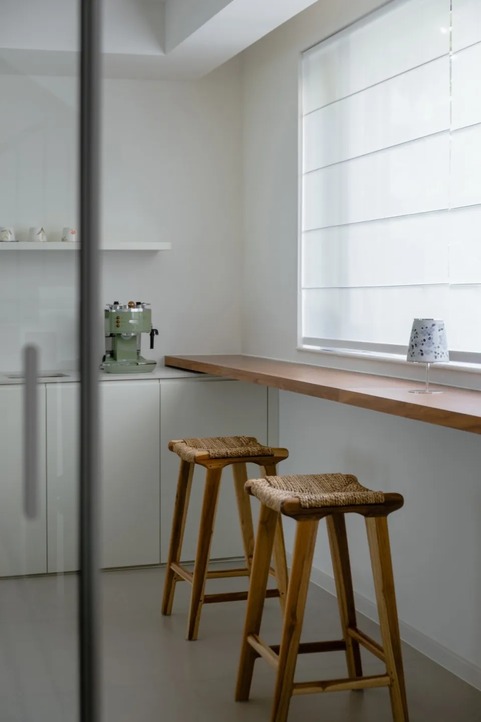 A modern minimalist kitchen with a wooden countertop and stools, featuring a green espresso machine and a window with blinds.
