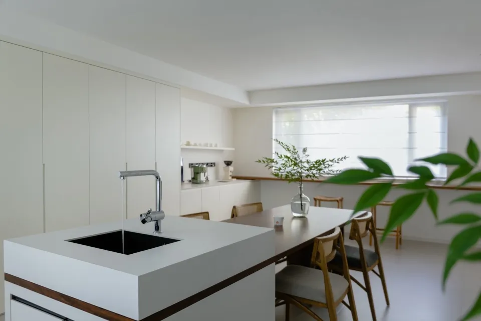 A modern kitchen with white cabinets, a white countertop, wooden table, and plants.