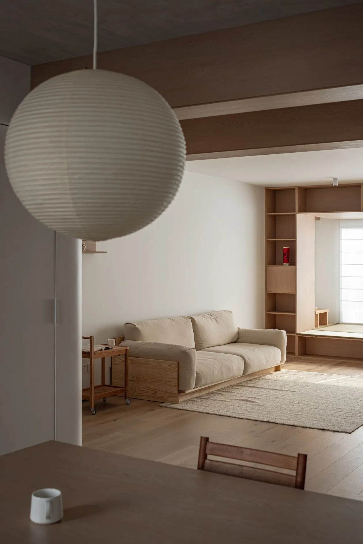 A modern minimalist living room featuring a wooden sofa, hanging lamp, and bookshelf.