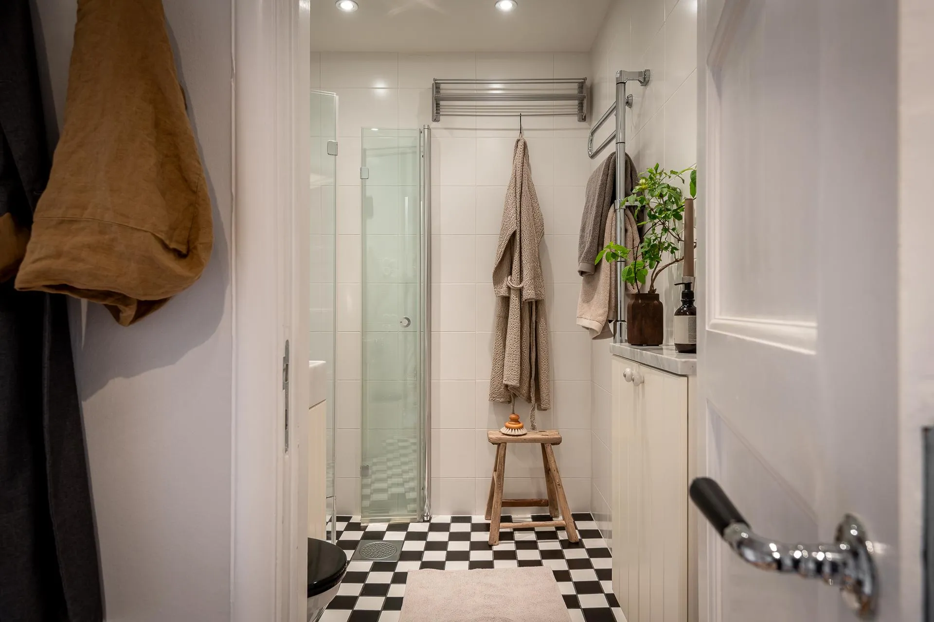 A minimalist bathroom featuring white tiled walls and floor, a glass shower stall, and hanging towels.