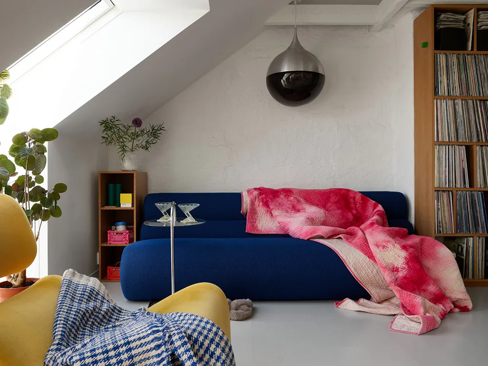 A modern minimalist living room features a blue sofa with a pink and red blanket, a yellow armchair with a blue and white bla