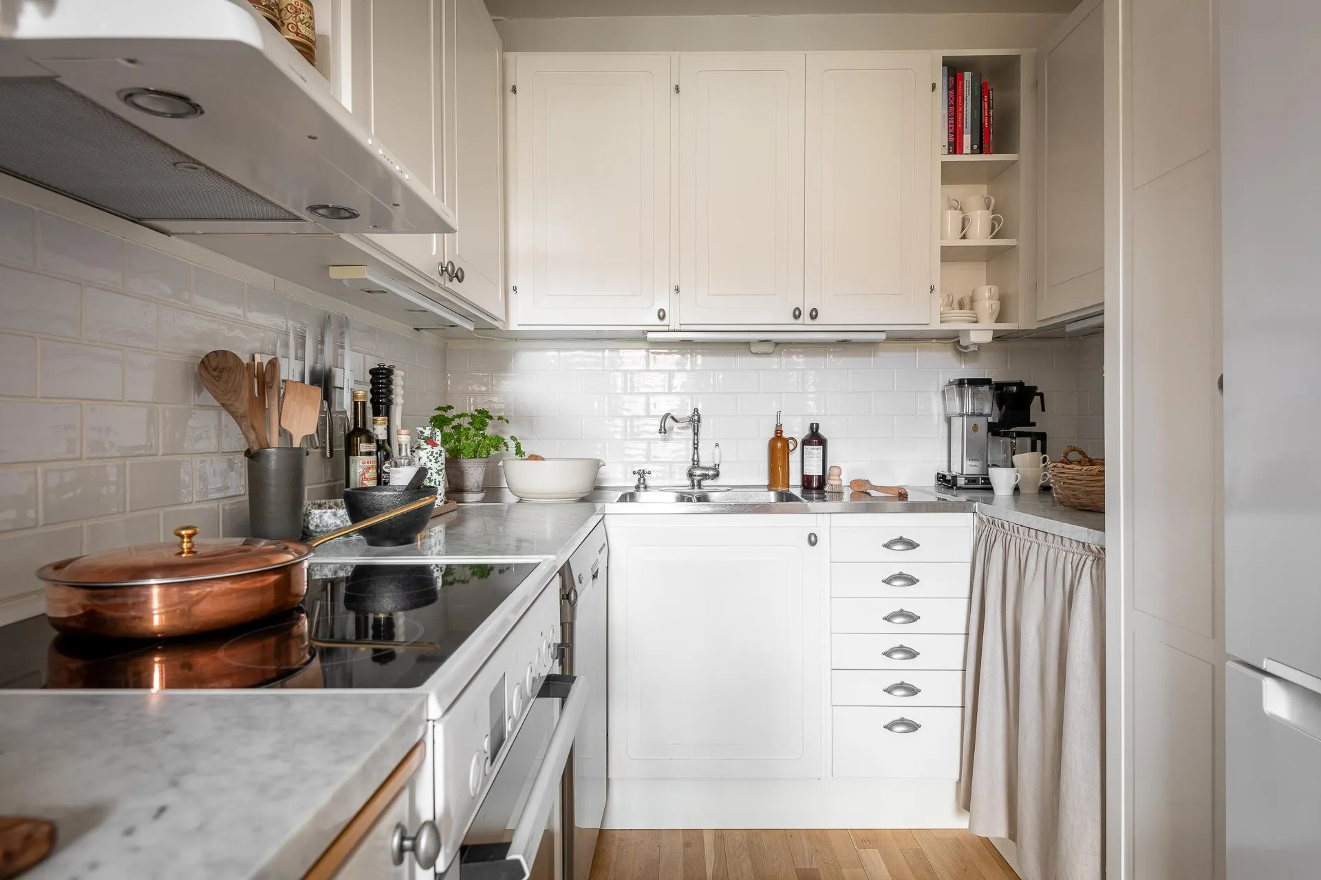 An image of a minimalist white kitchen with a white tile backsplash, stainless steel appliances, and light wooden flooring.