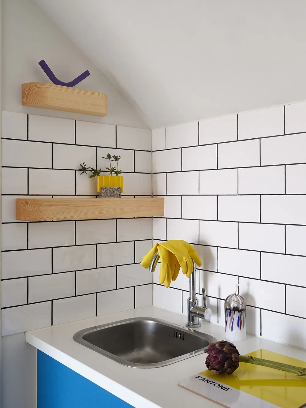 A minimalist kitchen with white ceramic tiles and a stainless steel sink, featuring a Pantone book and a yellow glove.