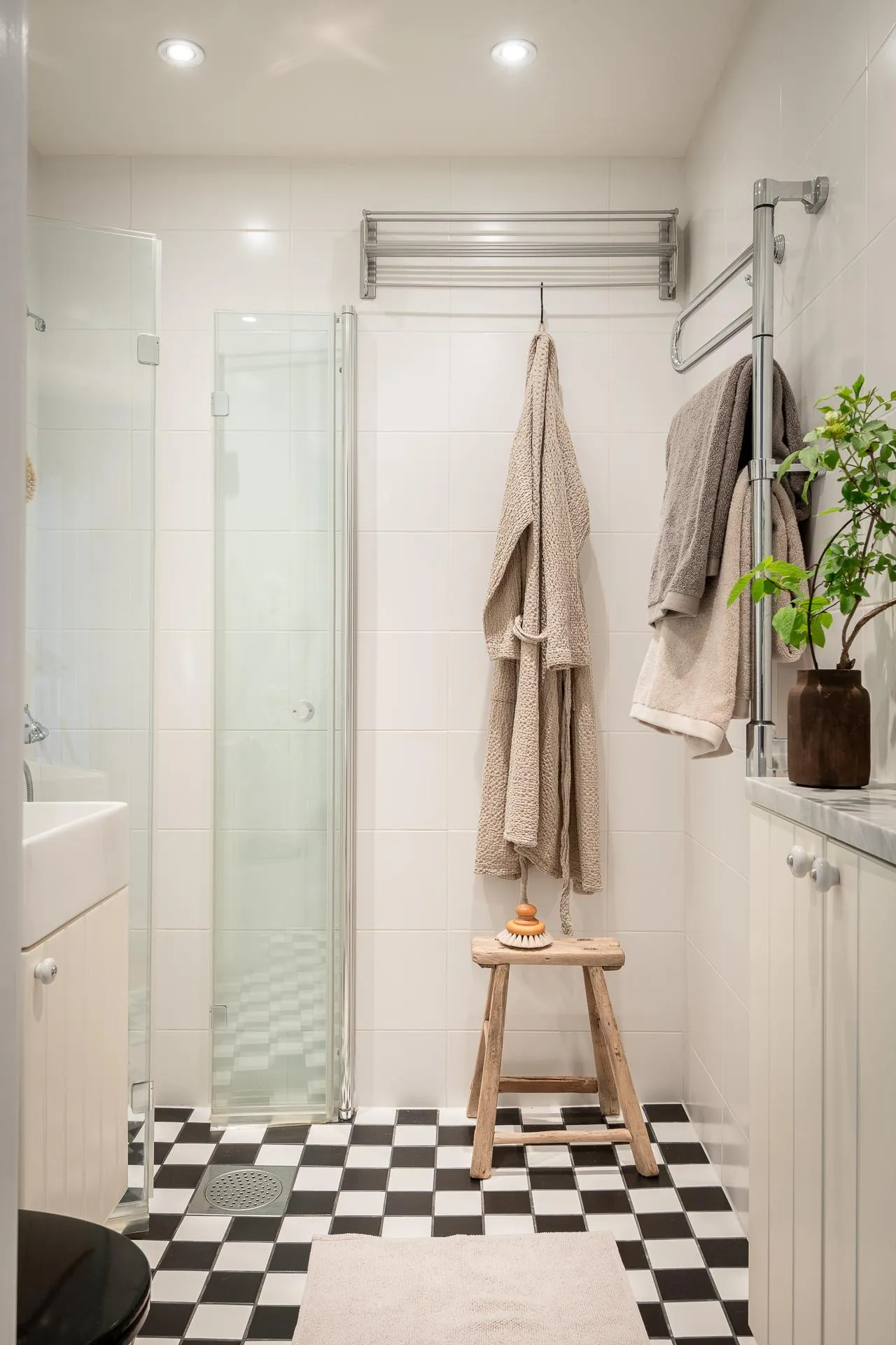 Modern bathroom interior with white tiled walls, checkered floor, glass shower enclosure, wooden stool, towel rack, and potte