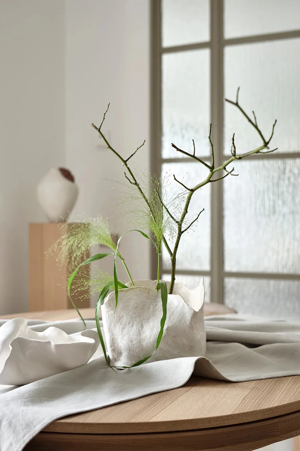 A modern minimalist white ceramic vase with green foliage and branches in a serene indoor setting.