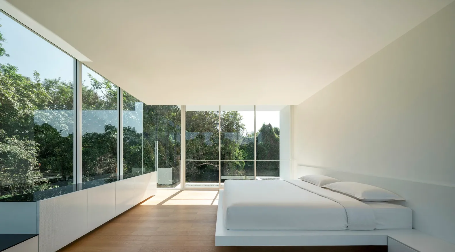 Minimalist bedroom with white furniture, large glass windows, and a view of green trees.