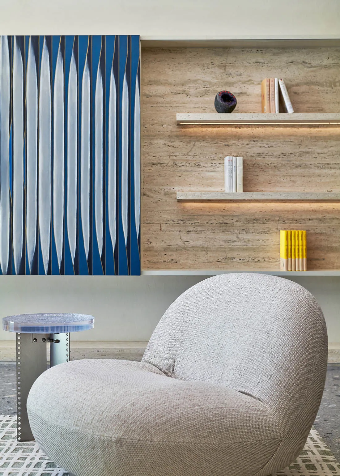 A minimalist living room with a gray upholstered chair, a transparent side table, and a blue and white abstract painting on a