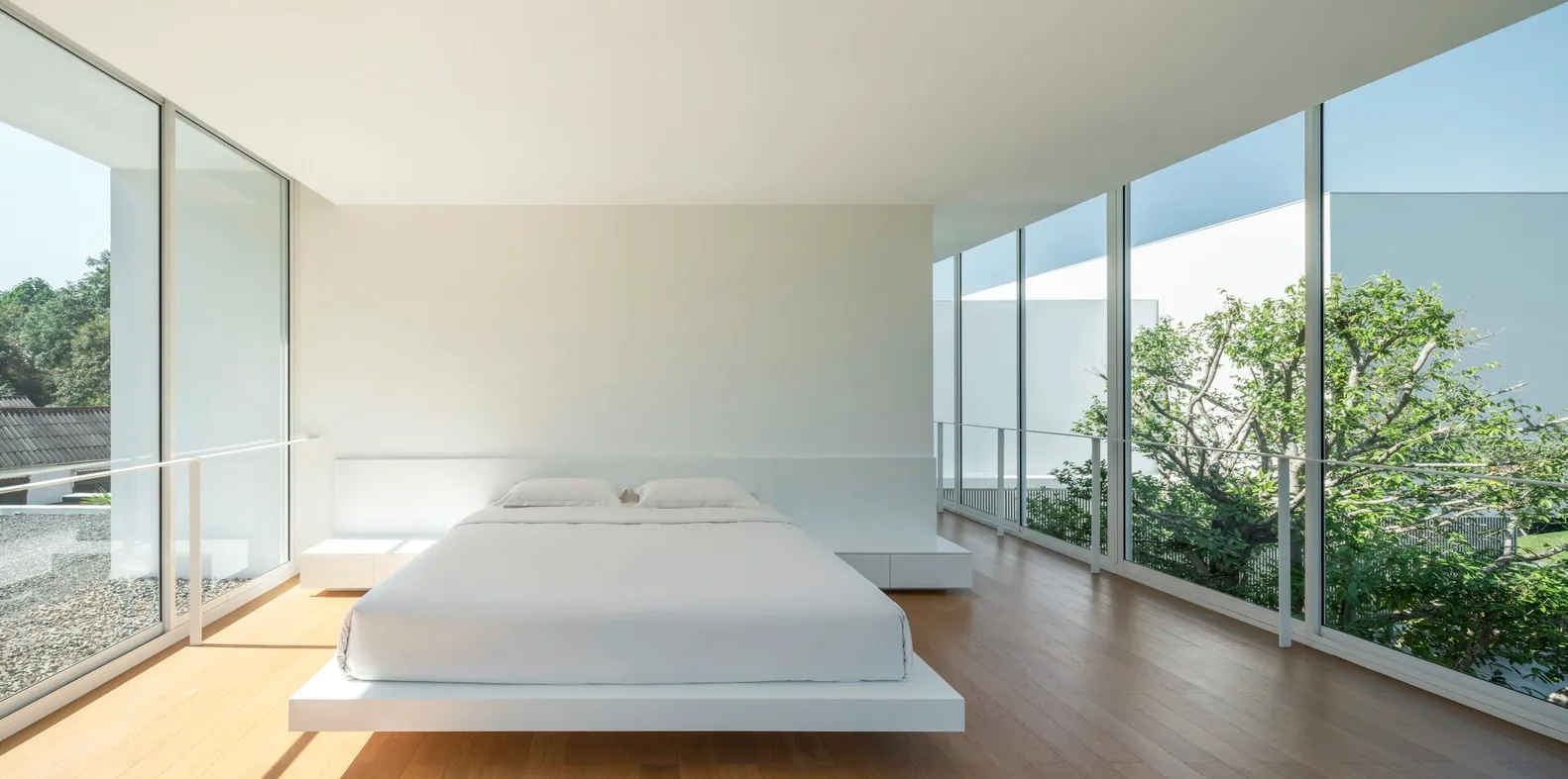 Minimalist bedroom with a single, white-floating platform bed with wooden flooring and large windows with a view of greenery