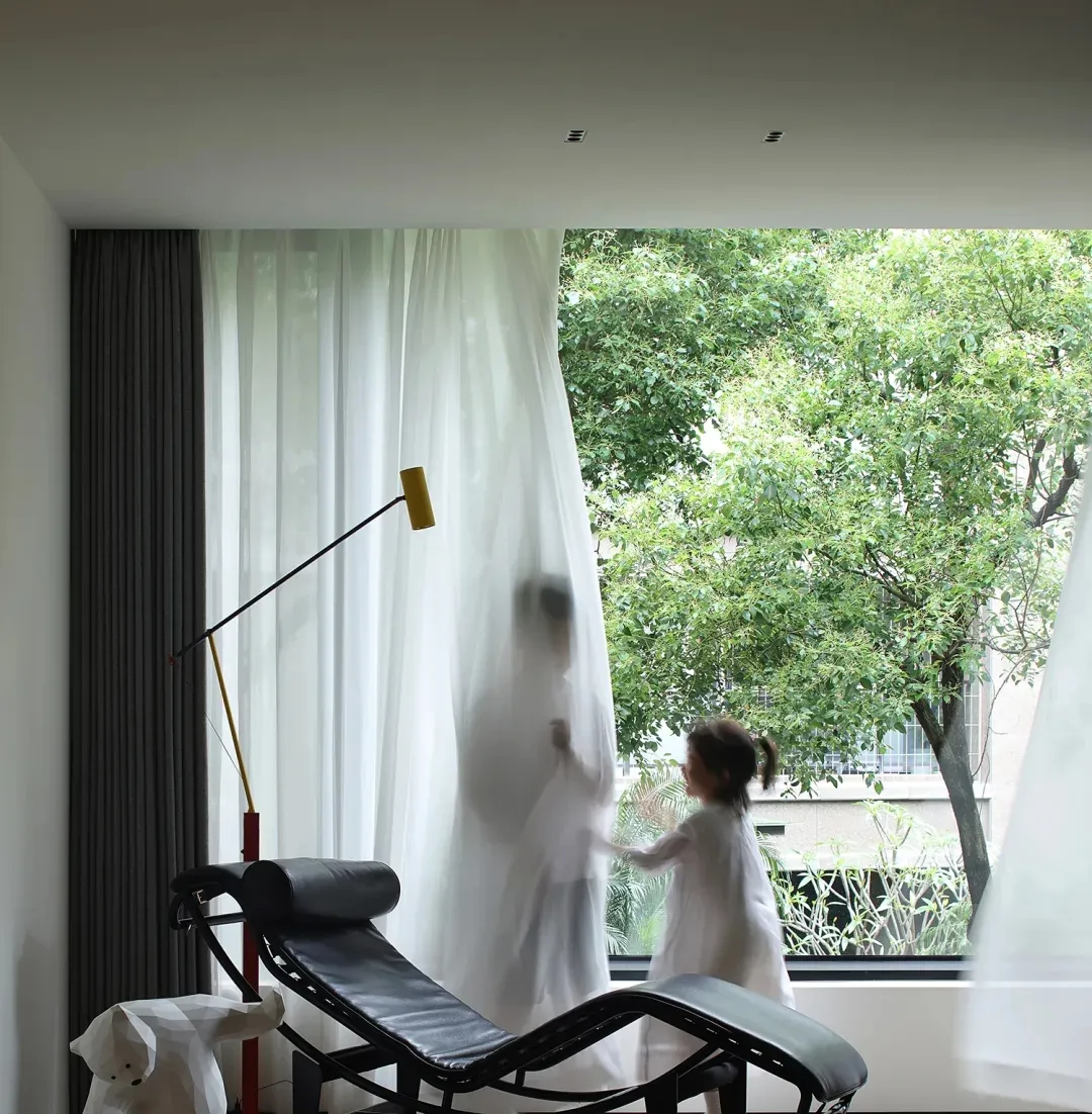 A contemporary living room featuring a sleek, black lounge chair with white curtains and a yellow desk lamp behind it, lookin