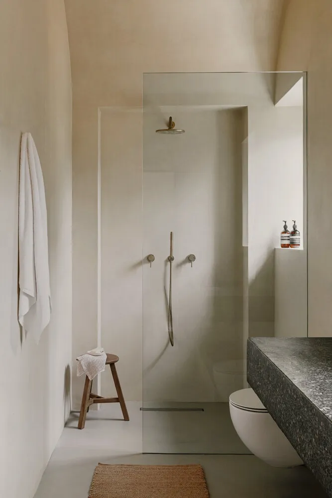 A minimalist concrete bathroom featuring a glass shower enclosure, stone sink, and wooden stool with a white towel.