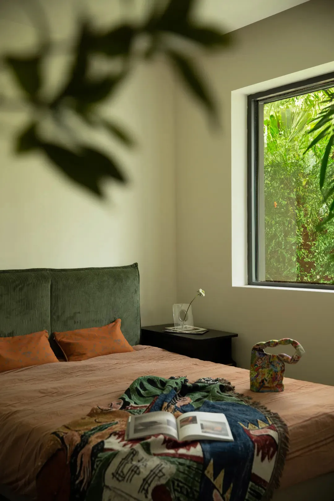 A modern bedroom featuring a bed with green headboard and colorful textiles, a small table with a vase and flower, and a wind