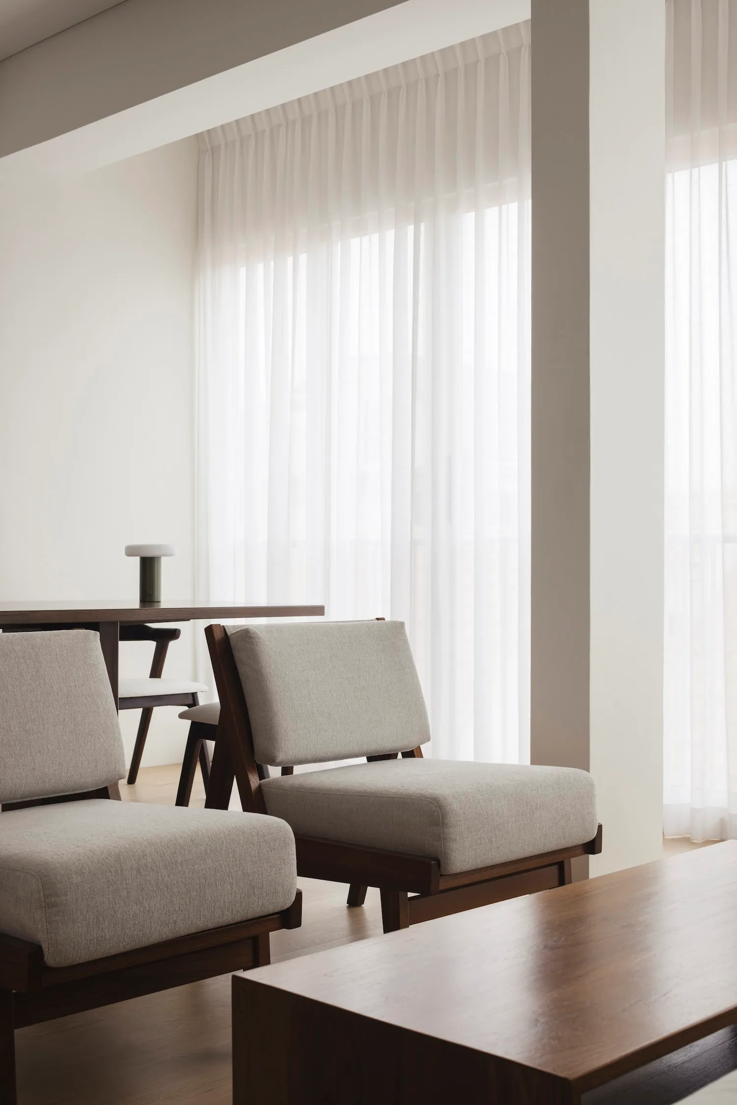 A modern minimalist living room featuring upholstered fabric chairs and wooden tables with sheer curtains allowing natural li