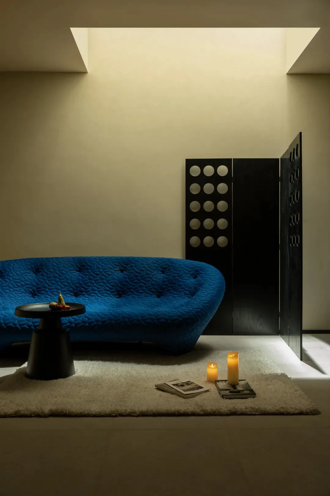 A minimalistic living room featuring a textured blue sofa, a small side table with a fruit bowl, and candles on a rug.