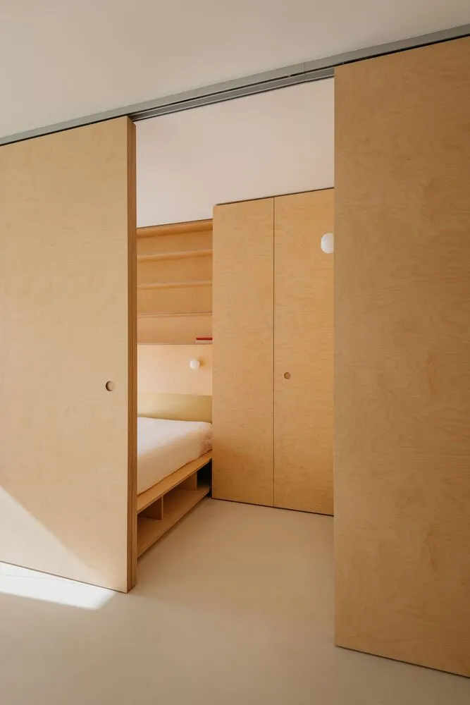 A minimalist bedroom with light wooden walls and paneling, featuring a bed with a storage base and a closet with sliding door