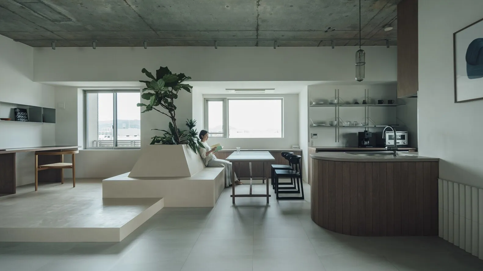 A modern kitchen with concrete and wooden finishes, featuring a plant on a elevated platform and a dining area with a woman r