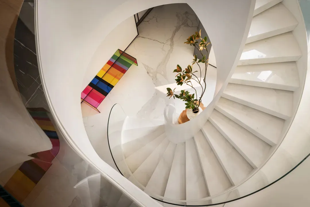 A white marble spiral staircase with glass railings and a potted plant.