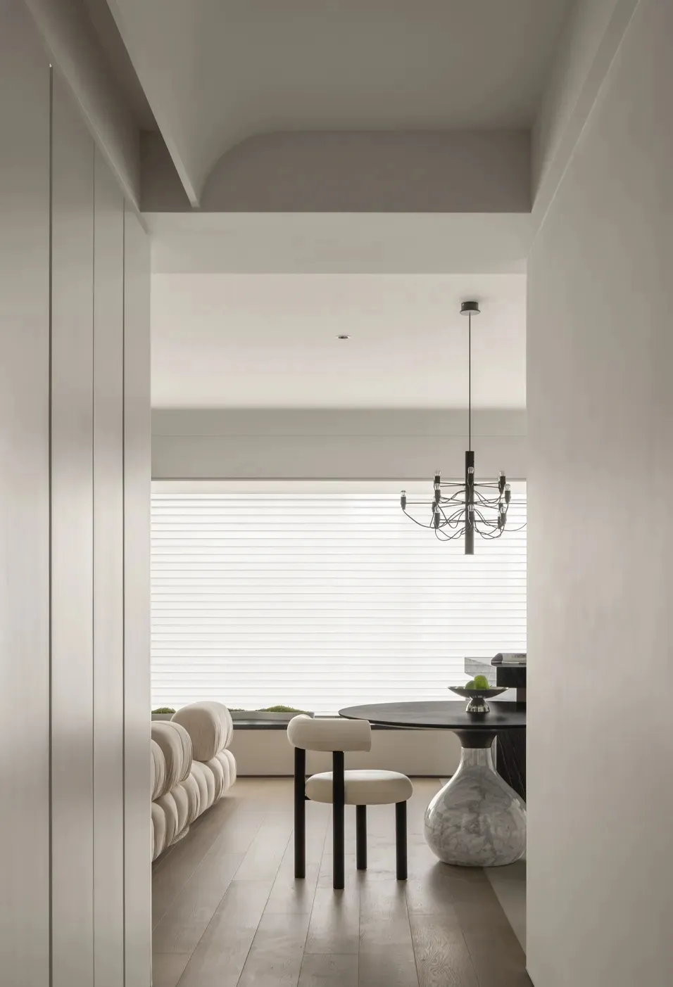 A modern living room with a white interior featuring a round black table, a sleek upholstered chair, and a minimalist chandel
