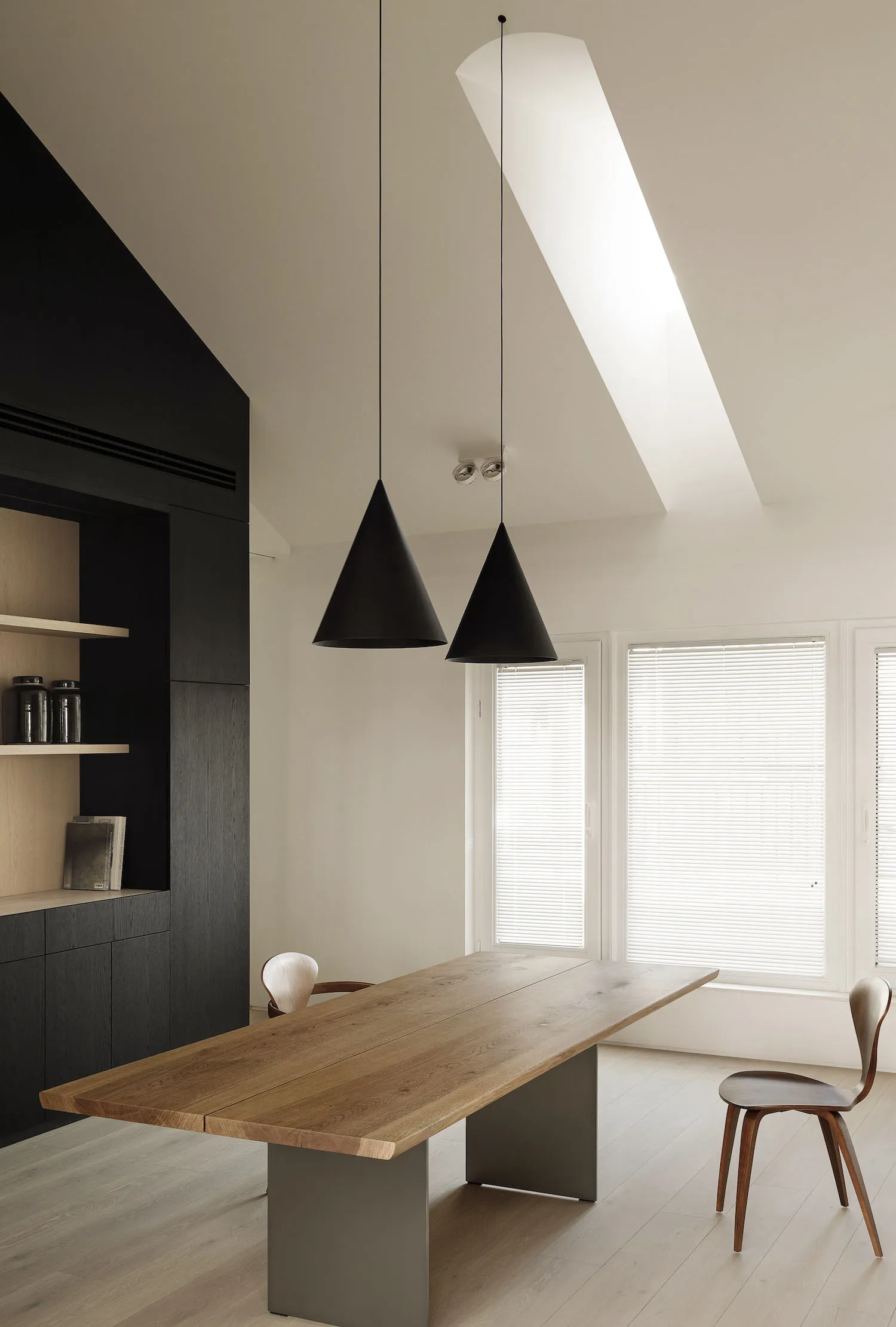 A minimalist dining room features a wooden table with metal legs, pendant lights, and a skylight.