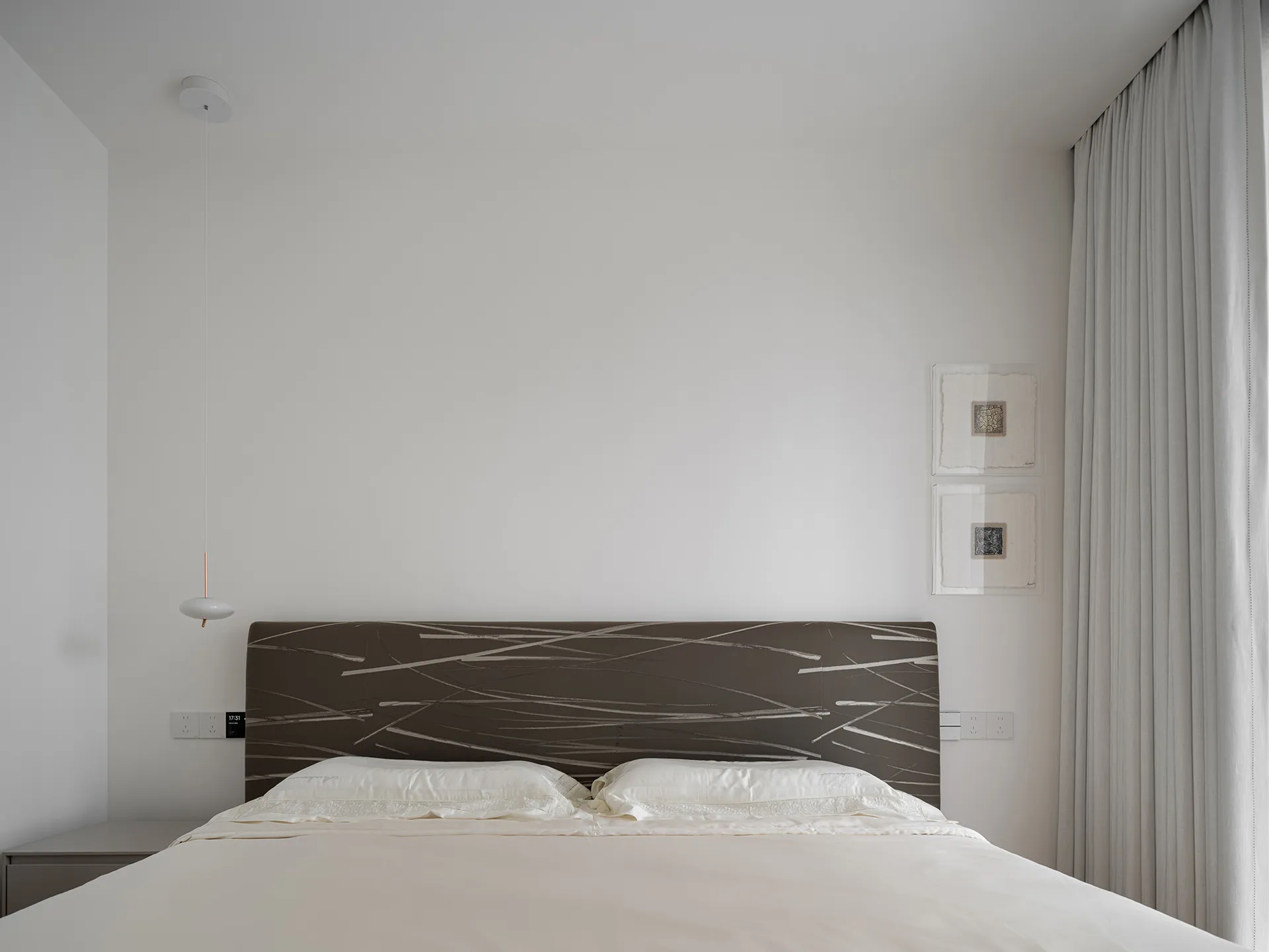 A minimalist bedroom featuring a bed with a patterned headboard and white bedding, alongside a white pendant light and a wind