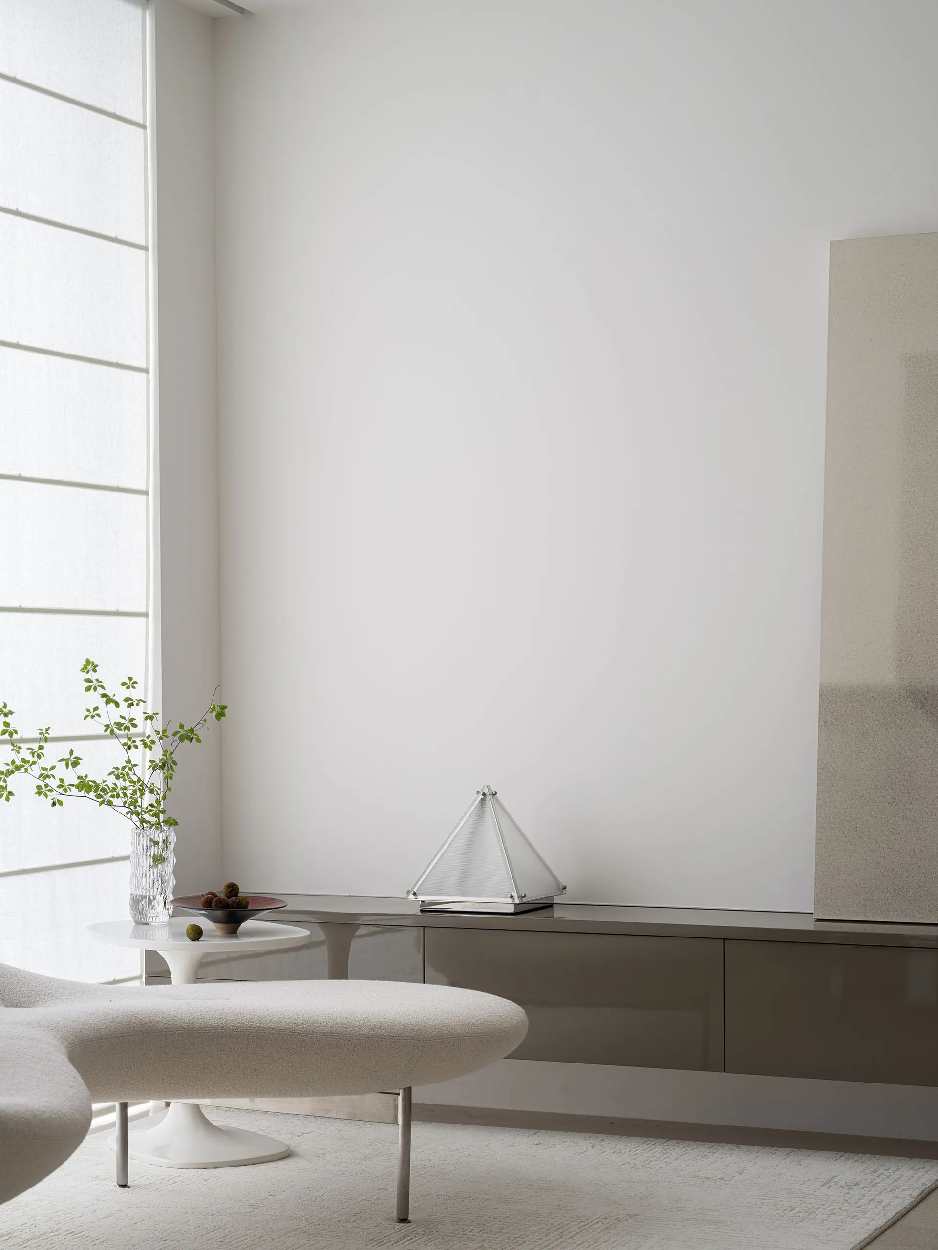 Modern minimalist living room featuring white furniture, a sleek glass cabinet, and a white ottoman with a white vase contain
