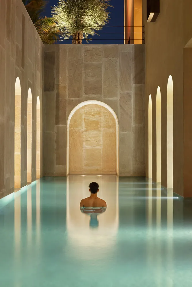 A serene swimming pool with a modern design and concrete walls, featuring a lone figure sitting in the pool.