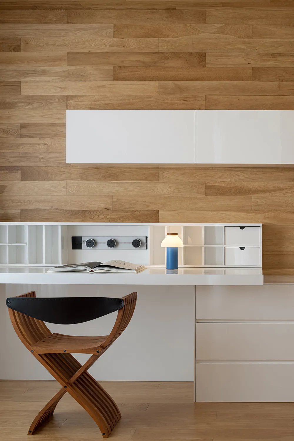 A modern home office featuring a wooden chair and a white desk with a blue lamp on it, set against a wooden wall with built-i