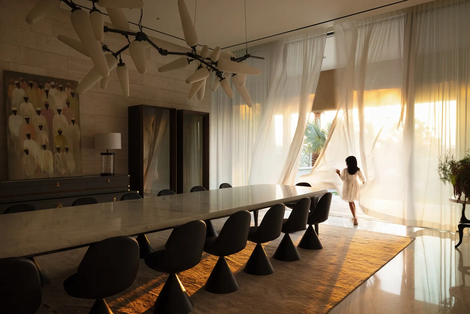 A modern living and dining room with a long marble table, glass chandelier, and sheer curtains.
