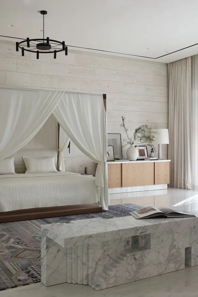 A modern minimalist bedroom featuring a canopy bed draped with white fabric, a marble bench, wooden drawers, and a textured r