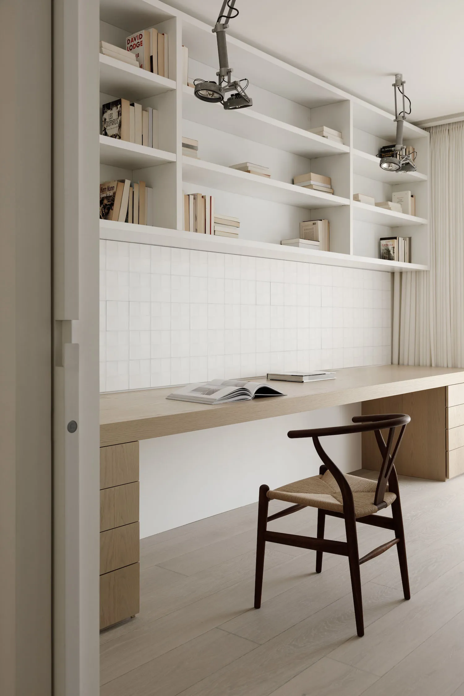 A minimalist home office with a long wooden desk, white bookshelves, and a woven chair.