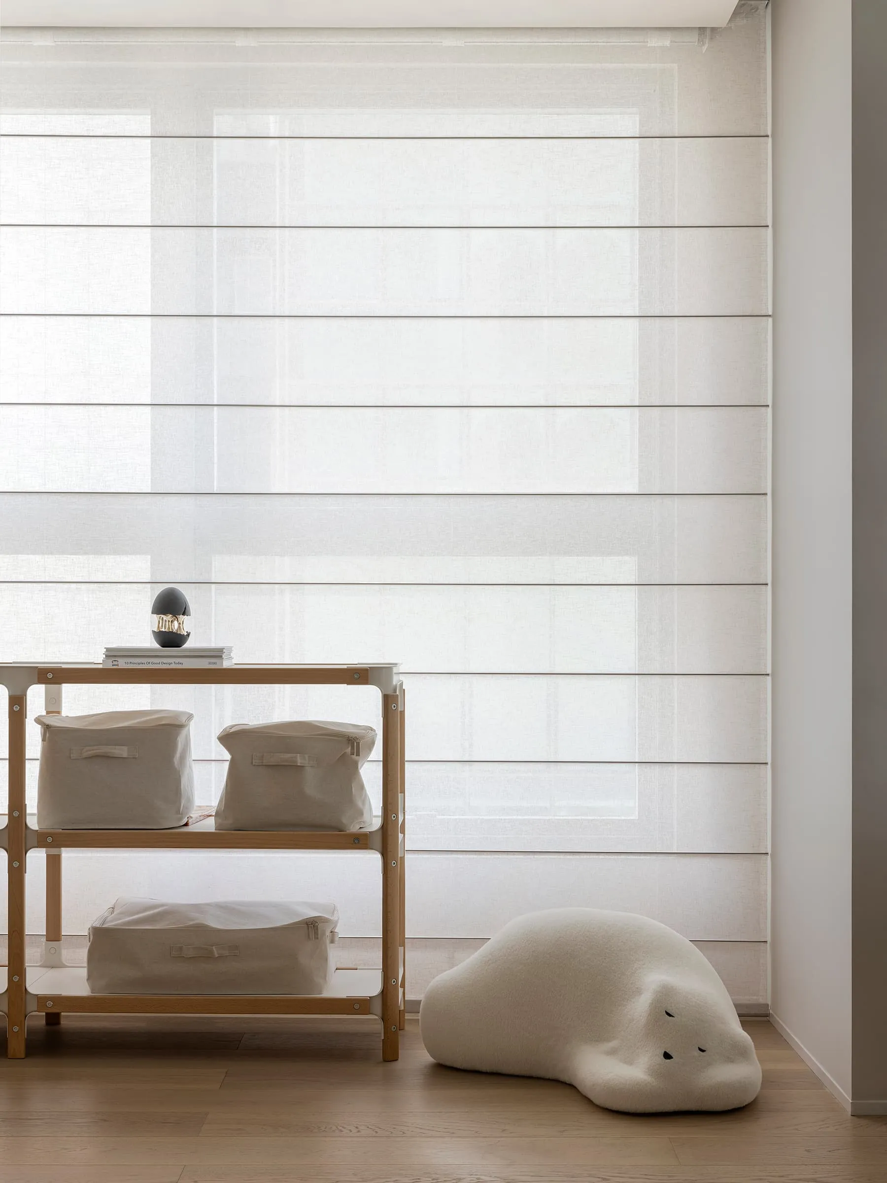 A minimalist interior featuring fabric blinds, storage bins, and a plush cloud-shaped cushion on a wooden floor.