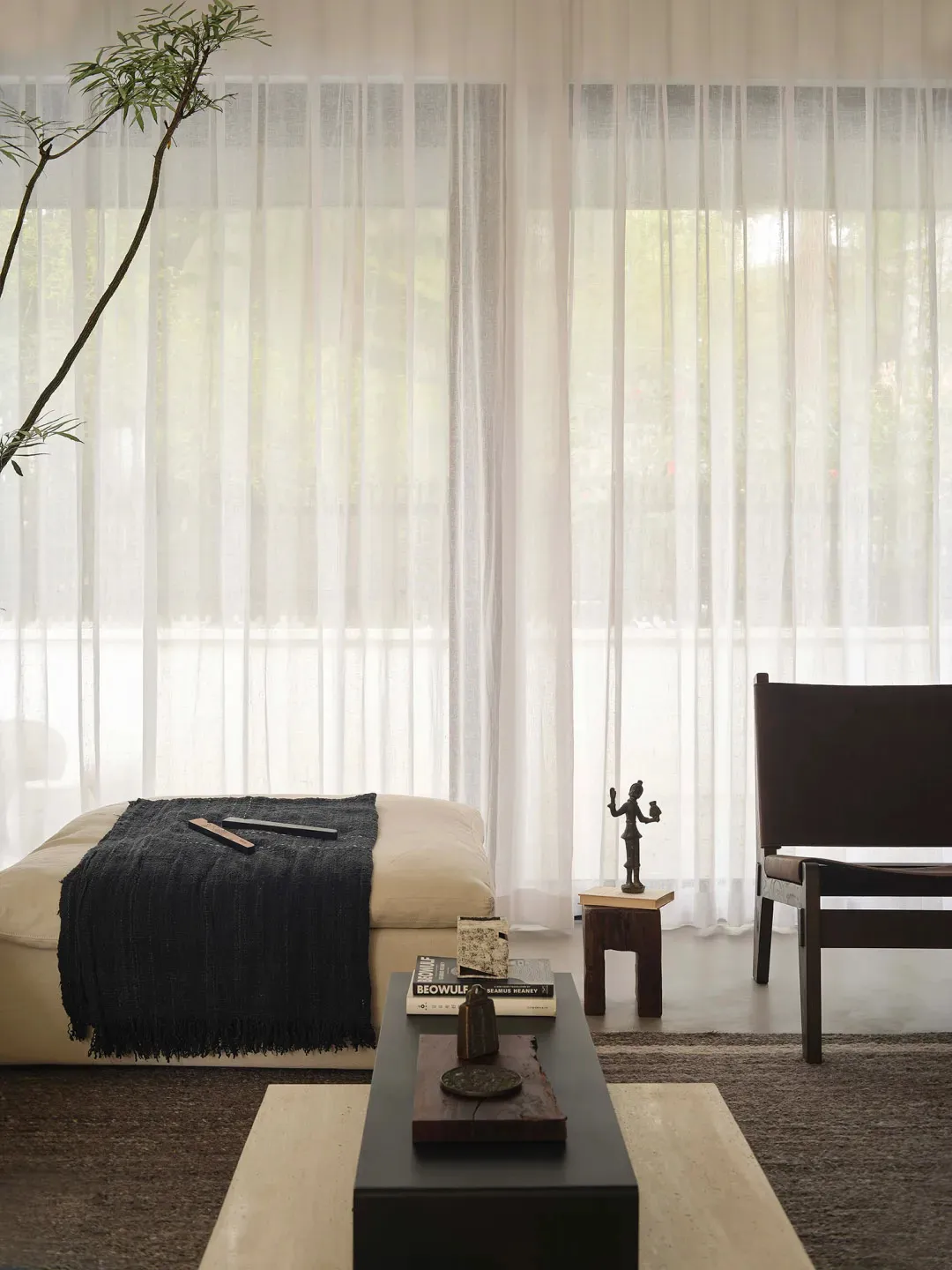 A minimalist living room with a beige sofa, black curtains, a black ottoman, and a black and wood coffee table.
