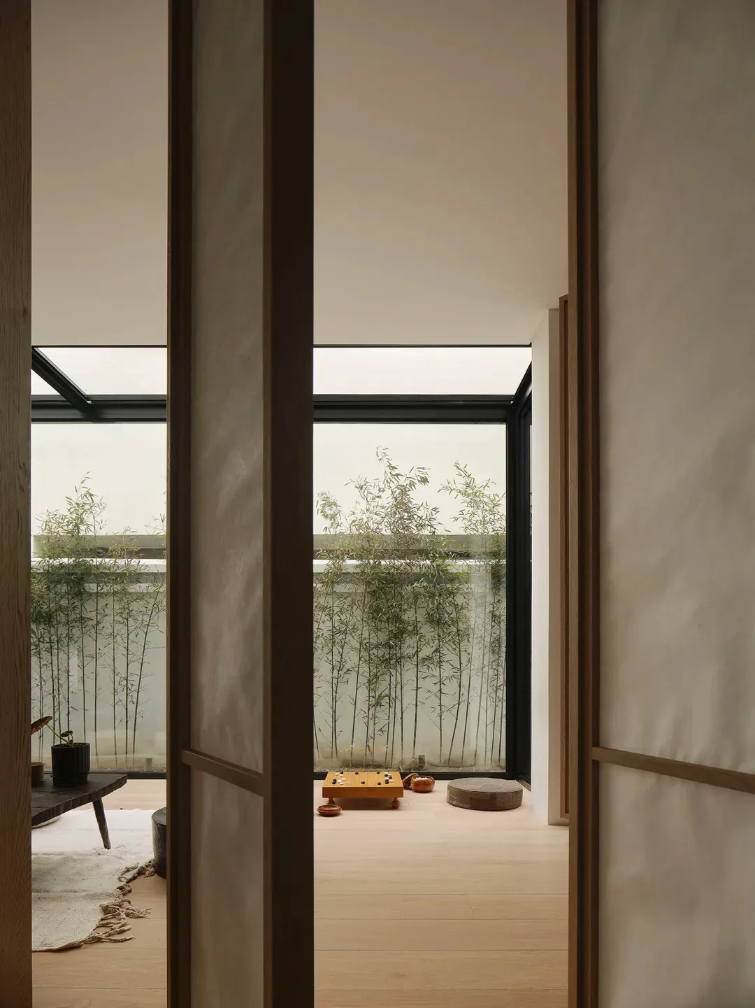 A minimalist living room with natural wood flooring and furnishings, featuring a bamboo garden outside the window.